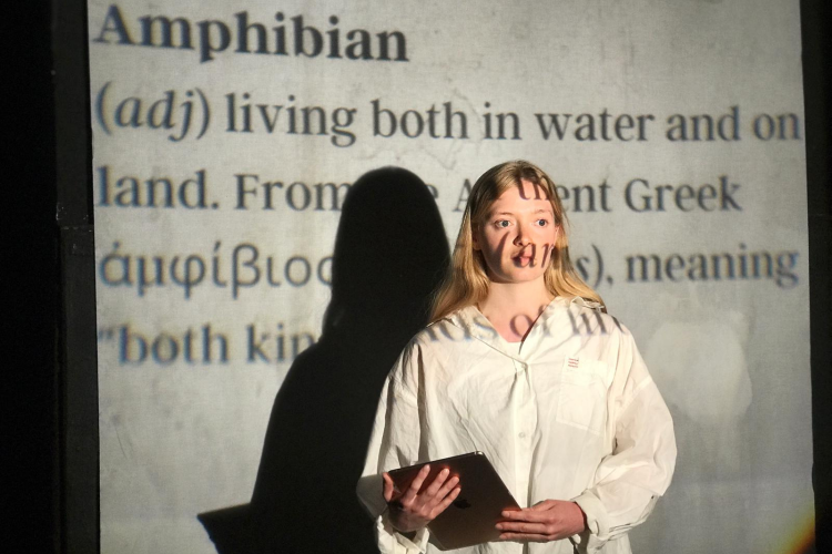 Actor holding a clipboard standing in front of a video projection of text giving the definition of 'Amphibian'